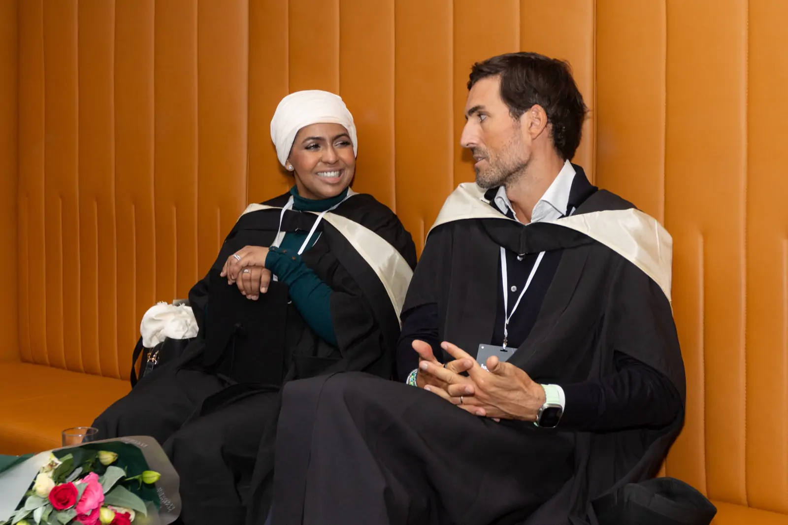 Ines Abdalla seated in conversation with a fellow graduate, both in robes